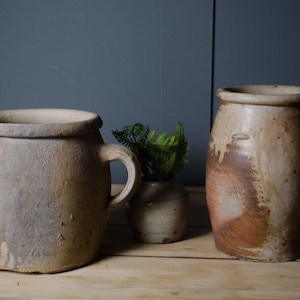 May include: Two rustic, wide-mouthed ceramic crocks with side handles and a smaller, central ceramic pot containing a green fern. The crocks feature a textured, mottled beige and brown finish with visible surface imperfections and earthy tones.