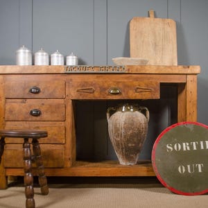 May include: A rustic wooden workbench with multiple drawers and a metal plaque reading JACQUES GIRARD. It features a wooden stool, four silver canisters, a wooden cutting board, a large ceramic vessel, and a round sign with the text SORTIE OUT.