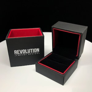 Puede incluir: Caja de anillo negra con interior rojo y el texto "REVOLUTION JEWELRY DESIGNS" impreso en la parte delantera.
