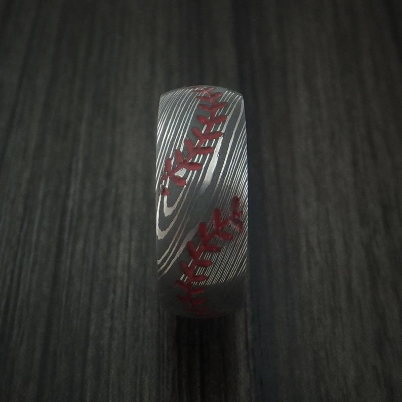 Puede incluir: Un anillo de plata con un dise&ntilde;o de puntadas de b&eacute;isbol. Las puntadas son rojas y el anillo tiene un acabado de metal cepillado.