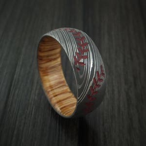 Puede incluir: Un anillo de plata y madera con un dise&ntilde;o de puntadas de b&eacute;isbol rojas. El anillo est&aacute; hecho de acero Damasco y tiene una incrustaci&oacute;n de madera.