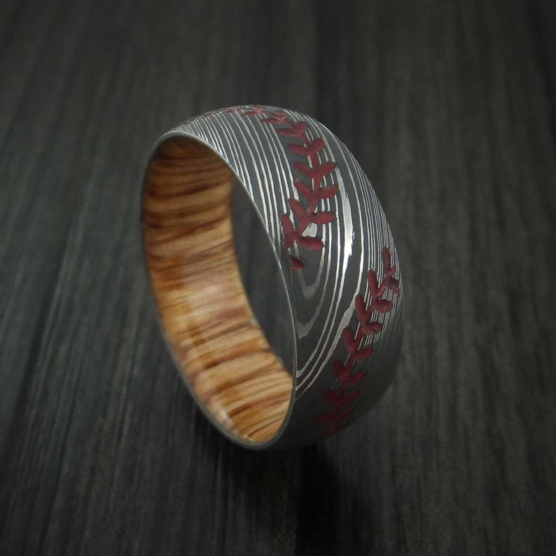 Puede incluir: Un anillo de plata y madera con un dise&ntilde;o de puntadas de b&eacute;isbol rojas. El anillo est&aacute; hecho de acero Damasco y tiene una incrustaci&oacute;n de madera.