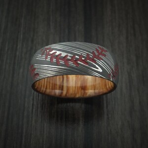 Puede incluir: Un anillo de b&eacute;isbol negro y rojo con incrustaciones de madera. El anillo est&aacute; hecho de acero damasco y presenta un dise&ntilde;o de costura roja.
