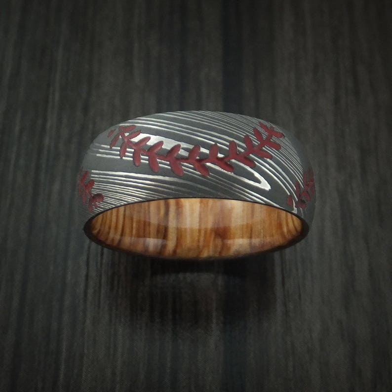 Puede incluir: Un anillo de b&eacute;isbol negro y rojo con incrustaciones de madera. El anillo est&aacute; hecho de acero damasco y presenta un dise&ntilde;o de costura roja.
