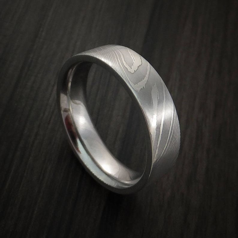 May include: A silver wedding band with a unique, patterned design. The band is made of metal and has a smooth, polished finish.
