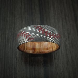 Puede incluir: Un anillo de plata y madera con un dise&ntilde;o de puntada de b&eacute;isbol. El anillo tiene un patr&oacute;n de puntada roja y una incrustaci&oacute;n de madera.