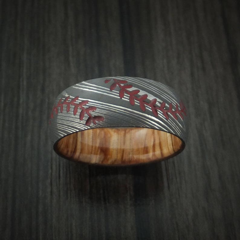 Puede incluir: Un anillo de plata y madera con un dise&ntilde;o de puntada de b&eacute;isbol. El anillo tiene un patr&oacute;n de puntada roja y una incrustaci&oacute;n de madera.