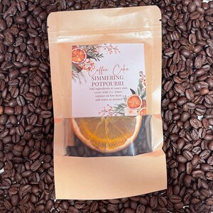 May include: A brown paper bag with a label that reads "Coffee Cake Simmering Potpourri". The label has a graphic of oranges, cinnamon sticks, and greenery. The bag contains dried orange slices and other ingredients for a simmering potpourri.