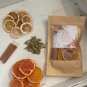 May include: A brown paper bag with a label that reads "Citrus Delight Simmering Potpourri". The bag contains dried orange slices, cinnamon sticks, and cardamom pods. The bag is sitting on a white surface with dried lemon slices and more cardamom pods.