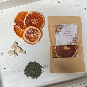 May include: Dried orange slices, ginger pieces, and a pile of dried green herbs are arranged on a white wooden surface. A kraft paper bag contains a grapefruit simmering potpourri mix. The bag has a label with the text "Meant to be Grapefruit Simmering Potpourri."