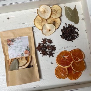 May include: A kraft paper bag labeled "Sweet Apple Pie Simmering Potpourri" with dried apple slices, star anise, cloves, orange slices, and bay leaves arranged on a white wooden surface. The potpourri ingredients are displayed for home fragrance.
