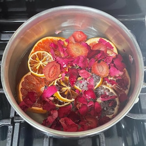 May include: A stainless steel pot filled with water, sliced oranges, lemons, strawberries, and red rose petals. The pot sits on a black stovetop, suggesting a simmering potpourri or herbal infusion.