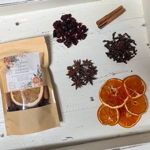 May include: A brown paper bag with a label that says "Welcome Home Simmering Potpourri". The bag contains dried orange slices, cinnamon sticks, cloves, star anise, and cranberries.