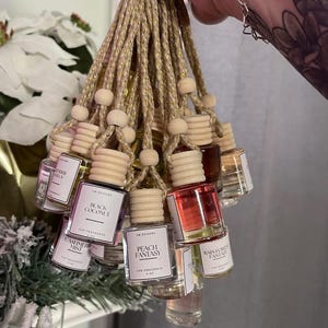 May include: A collection of car air fresheners in small glass bottles with wooden caps, hanging from gold and beige cords. The labels display various scents, including "Black Coconut" and "Peach Fantasy". The bottles are held in a hand.