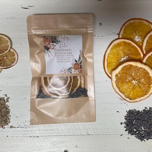 May include: A brown paper bag with a label that reads "Relaxing Simmering Potpourri" and contains dried orange slices, lavender buds, and dried herbs. The bag is sitting on a white wooden surface with dried orange slices, lavender buds, and dried herbs scattered around it.