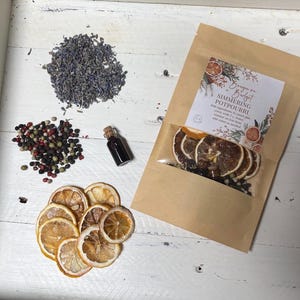May include: A brown paper bag with a clear window showing dried orange slices, peppercorns, and lavender. The bag has a label that reads "Bougie on a Budget Simmering Potpourri". The label also includes instructions for use.  The bag is on a white surface with a small bottle of essential oil, dried lavender, and peppercorns.