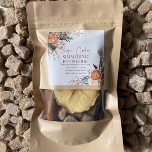 May include: A brown paper pouch with a clear window showing dried fruit, cinnamon sticks, and vanilla beans. The pouch has a label that reads "Sugar Cookie Simmering Potpourri" and instructions for use.