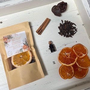 May include: A brown paper bag with a label that reads "Vanilla Orange Dreamcicle Simmering Potpourri". The bag contains dried orange slices, cinnamon sticks, and a small bottle of essential oil. There are also dried orange slices, cloves, and dried plums on a white surface.
