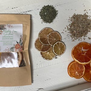May include: A brown paper bag with a label that reads "Eucalyptus Lime Sunshine Simmering Potpourri". The bag contains dried citrus fruit and herbs. The image also shows dried citrus fruit and herbs on a white surface.