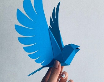 Peut inclure: Un oiseau en papier bleu avec des ailes pliées, réalisé dans un style origami 3D. L'oiseau est tenu dans une main avec des ongles peints.