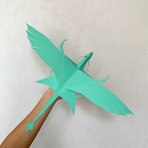 May include: A teal paper dragon with wings outstretched and a long tail. The dragon is held by a hand.