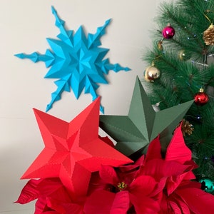 May include: A red paper star, a green paper star, and a blue paper snowflake are displayed in front of a potted poinsettia plant. The poinsettia plant has red leaves and is in a brown pot. The paper stars and snowflake are decorations for the holiday season.