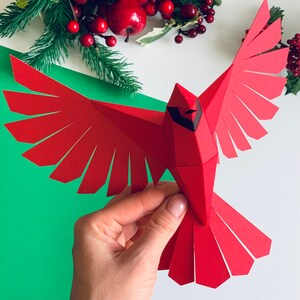 Pu&ograve; includere: Un uccello cardinale di carta rosso con occhi e becco neri, piegato in un design di origami 3D. L'uccello &egrave; tenuto in una mano su uno sfondo verde.