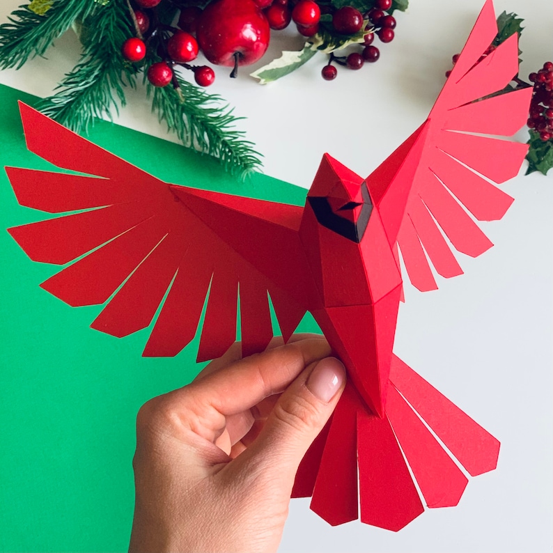 Pu&ograve; includere: Un uccello cardinale di carta rosso con occhi e becco neri, piegato in un design di origami 3D. L'uccello &egrave; tenuto in una mano su uno sfondo verde.