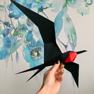 以下が含まれることがあります： 赤いアクセントのある黒い折り紙の鳥。鳥は紙でできており、リアルな形に折り畳まれています。鳥は手で持たれています。