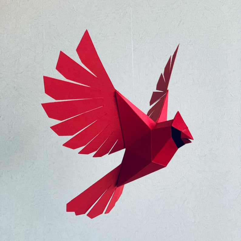 Pu&ograve; includere: Un mobile di carta rosso a forma di cardinale con le ali spiegate in volo. L'uccello &egrave; fatto di carta piegata e ha un design geometrico.