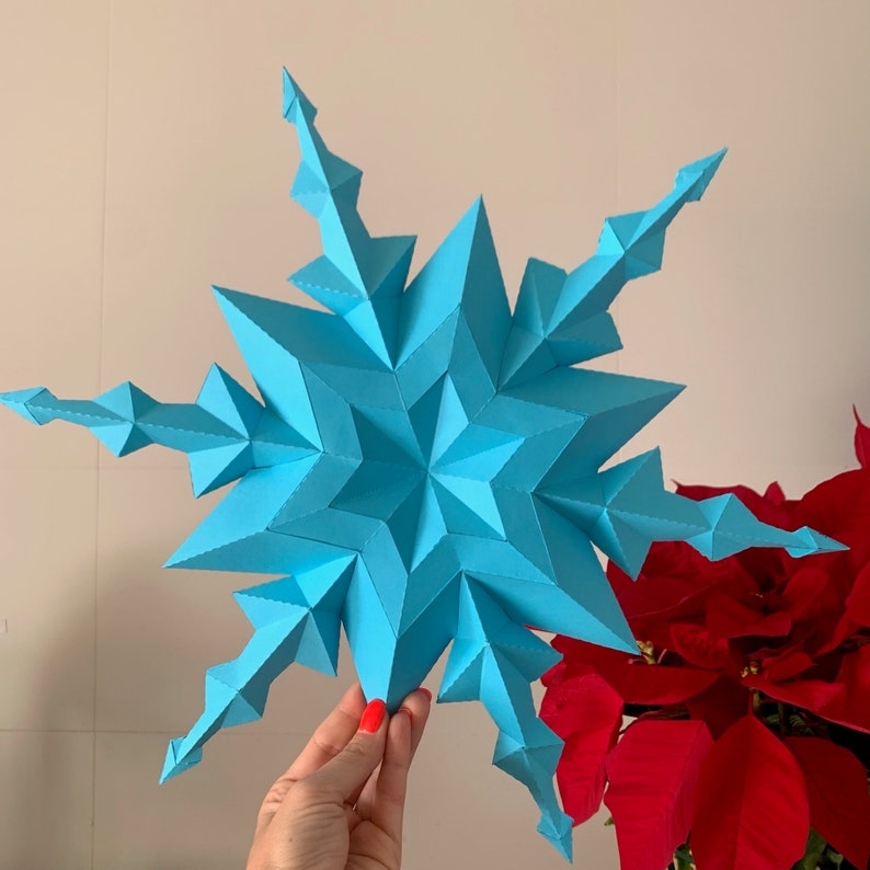 May include: A large, blue paper snowflake with a 3D design. The snowflake is held in a hand against a white background.