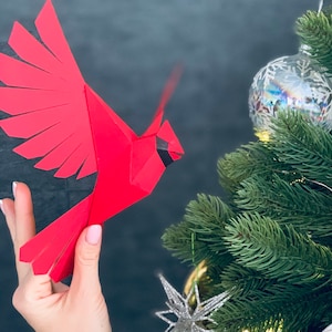 Pu&ograve; includere: Un cardinale di origami rosso con occhi e becco neri, tenuto in una mano su uno sfondo grigio scuro. Un albero di Natale con aghi verdi e un ornamento argentato &egrave; sullo sfondo.