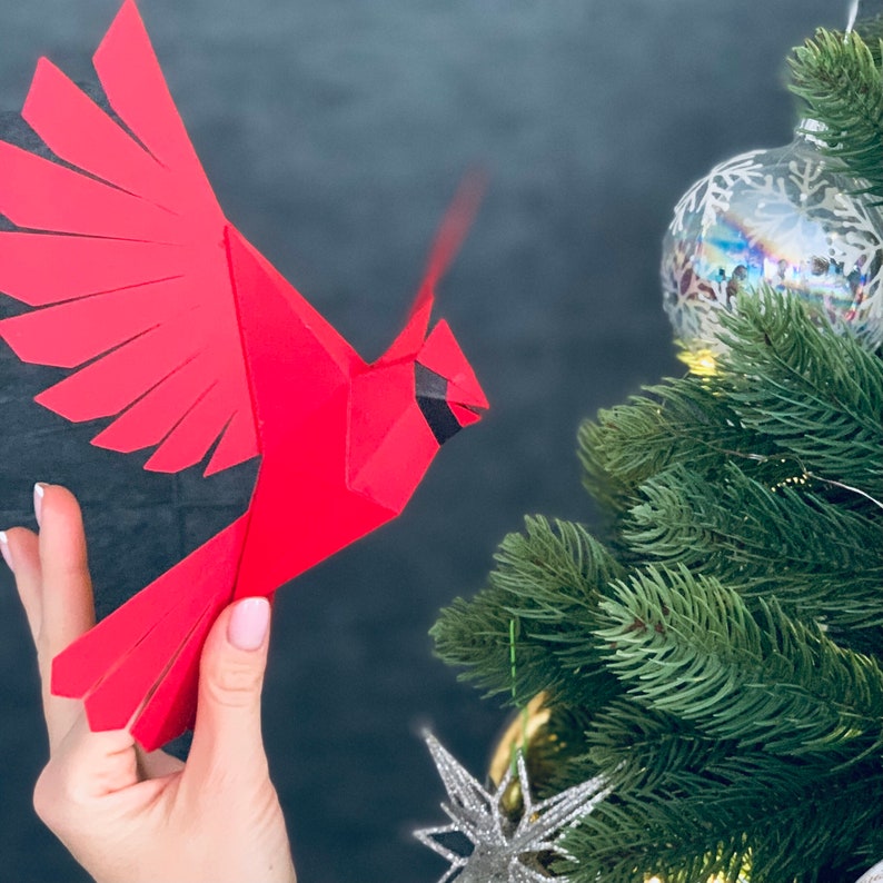 Pu&ograve; includere: Un cardinale di origami rosso con occhi e becco neri, tenuto in una mano su uno sfondo grigio scuro. Un albero di Natale con aghi verdi e un ornamento argentato &egrave; sullo sfondo.