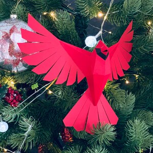 Pu&ograve; includere: Un ornamento di carta rosso a forma di cardinale con becco e occhi neri, appeso a un albero di Natale verde.