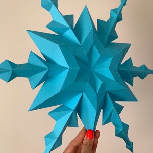 May include: A large, blue, paper snowflake with a 3D design. The snowflake is made of many small triangles and has a starburst shape.