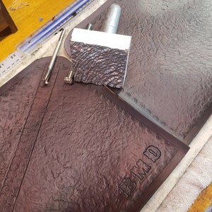 May include: A brown leather piece with a textured surface and the letters "BMMD" embossed on it. The leather is being worked on with tools and a metal stamp.