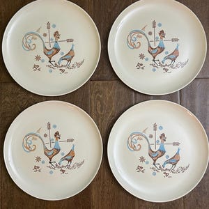 May include: A set of four round, off-white ceramic dinner plates, each featuring an identical central illustration of a rooster and a smaller bird in shades of brown and light blue, accented with small floral and wheat-like graphic motifs.