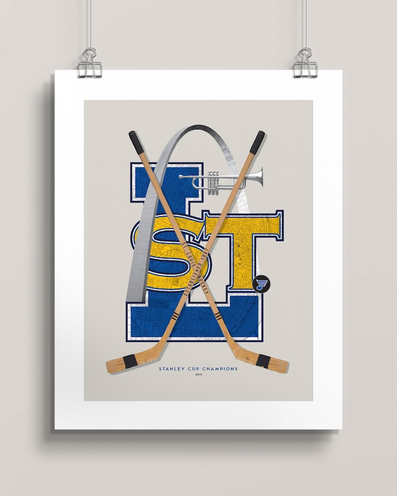 May include: A blue and yellow graphic with the letters "STL" and a silver archway. Two hockey sticks are crossed behind the letters. The text "STANLEY CUP CHAMPIONS 2019" is below the graphic.