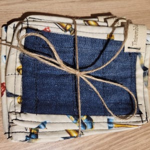 May include: A stack of square fabric coasters tied with twine. The coasters have a denim blue center and a patterned border with a cream background and colorful illustrations. The tag reads "Thimmble".