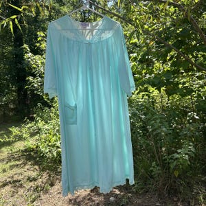 May include: A light mint green button-down robe featuring short sleeves, a V-neckline with delicate lace trim, and a single patch pocket on the front. The garment is made of a smooth, lightweight fabric and hangs from a white plastic hanger.