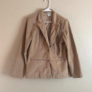 May include: A tan corduroy blazer with a notched lapel, two front pockets, and a two-button closure. The jacket is hanging on a white hanger against a neutral background. The blazer is a classic style.