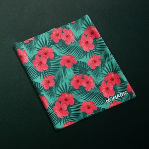 Puede incluir: Una tela verde con un estampado tropical con flores de hibisco rosas y hojas verdes. La tela tiene la palabra "NOMADIC" impresa en blanco en la esquina inferior derecha.