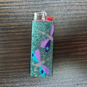 May include: A rectangular lighter encased in a beaded sleeve featuring a repeating dragonfly pattern. The beads are primarily metallic green, with purple, light blue, and silver accents forming the insect shapes against the textured surface.