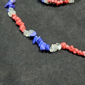 May include: A necklace made of red, white, and blue gemstone chips. The necklace is made of small, irregular shaped chips of red, white, and blue gemstones. The necklace is on a black background.
