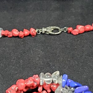 May include: A red, white, and blue gemstone necklace with a silver clasp. The necklace is made of small, irregular shaped stones.