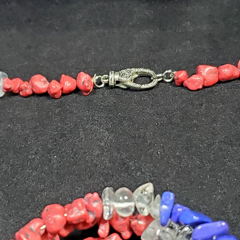 May include: A red, white, and blue gemstone necklace with a silver clasp. The necklace is made of small, irregular shaped stones.