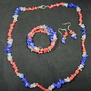 May include: A set of red, white, and blue gemstone jewelry, including a necklace, bracelet, and earrings. The necklace and bracelet are made of small, irregular shaped stones, while the earrings are teardrop shaped.