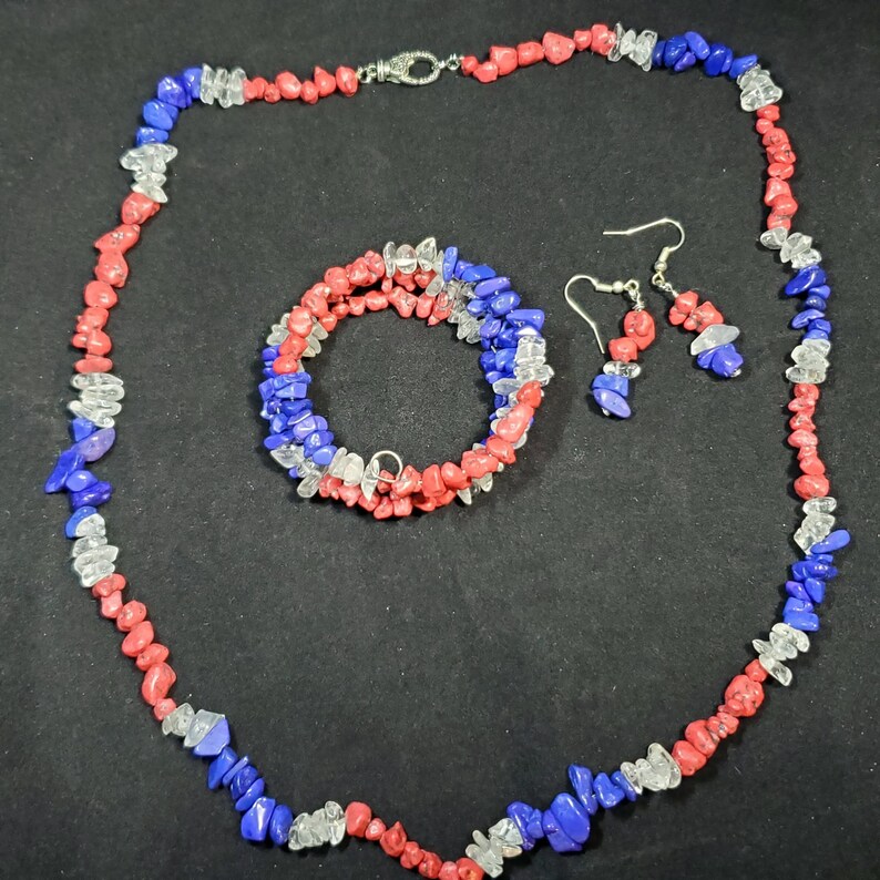 May include: A set of red, white, and blue gemstone jewelry, including a necklace, bracelet, and earrings. The necklace and bracelet are made of small, irregular shaped stones, while the earrings are teardrop shaped.