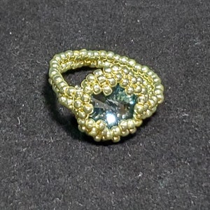 May include: A gold beaded ring featuring a large, faceted, teal gemstone in the centre.