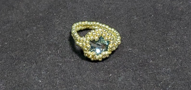 May include: A gold beaded ring featuring a large, faceted, teal gemstone in the centre.